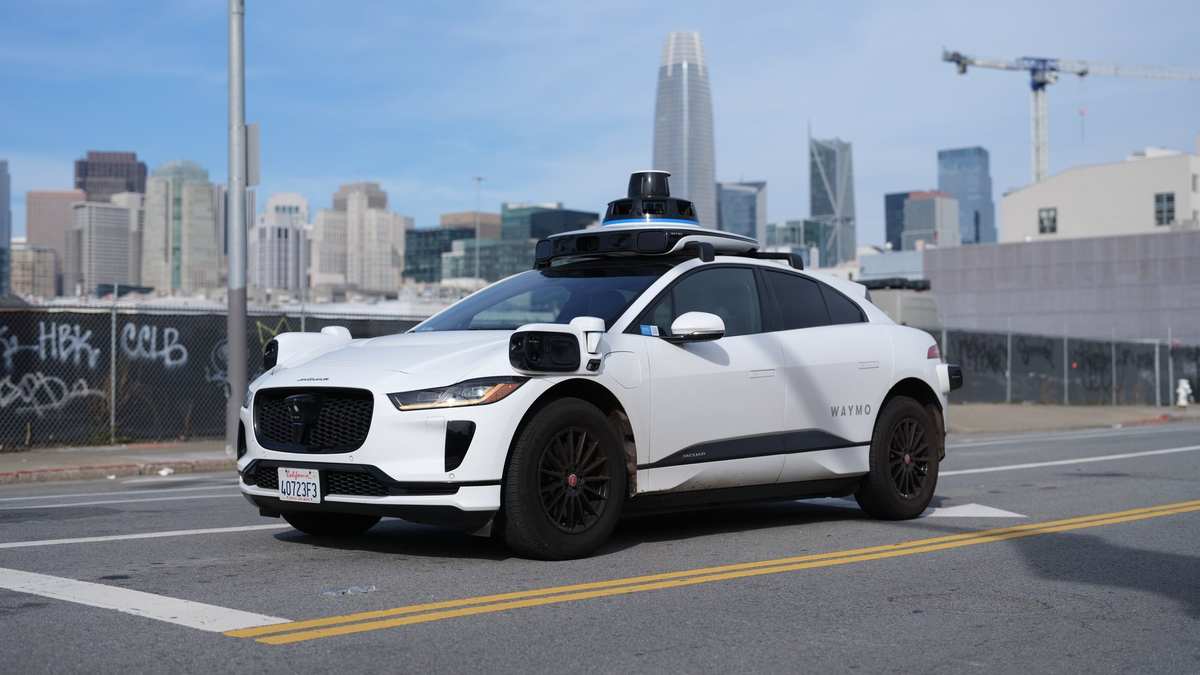 White Waymo Jaguar I-Pace with nobody at the wheel on a San Francisco street, with the city's skyscrapers in the background